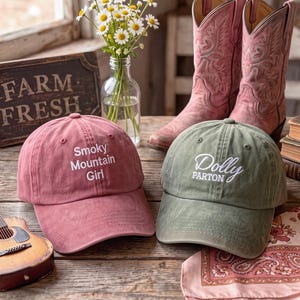 Op de afbeelding: Een oudroze baseballpet met de witte geborduurde tekst Smoky Mountain Girl en een olijfgroene baseballpet met de witte geborduurde tekst Dolly Parton, beide voorzien van een gebogen klep en een gestructureerde kroon met ventilatiegaatjes.