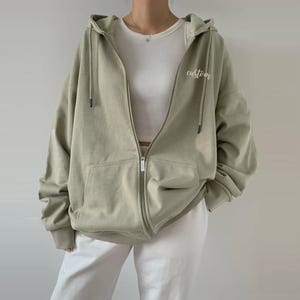 Op de afbeelding: Een lichtgroen hooded sweatshirt met een ritssluiting. Het sweatshirt heeft een zak aan de voorkant en het woord "custom" geborduurd aan de rechterkant.