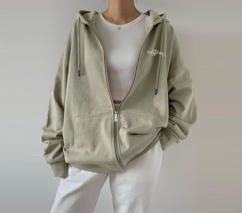 Op de afbeelding: Een lichtgroen hooded sweatshirt met een ritssluiting. Het sweatshirt heeft een zak aan de voorkant en het woord "custom" geborduurd aan de rechterkant.