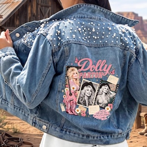 May include: A light blue denim jacket featuring white pearl embellishments across the upper back and shoulders. The back panel displays a graphic print of Dolly Parton with a guitar, pink cowboy boots, flowers, butterflies, and the text Dolly Parton.