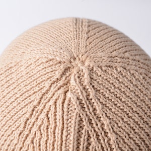 May include: Close-up of a beige knitted beanie hat. The hat is made of a soft, textured yarn and has a simple, classic design.