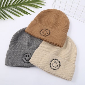 May include: Three knit beanies in brown, gray, and beige with a black embroidered smiley face and the text "smile smile smile" around the face.