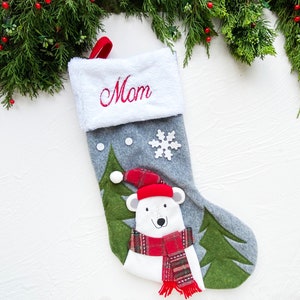 May include: A gray Christmas stocking with a white cuff embroidered with the word "Mom" in red. The stocking features a polar bear wearing a red hat and scarf, and green felt trees. A white snowflake and two white dots are also on the stocking.