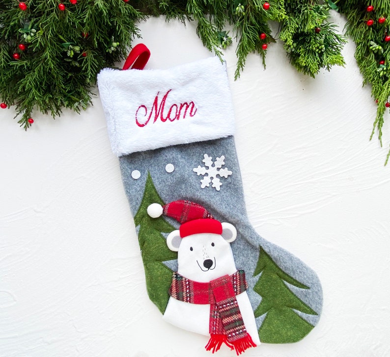 May include: A gray Christmas stocking with a white cuff embroidered with the word "Mom" in red. The stocking features a polar bear wearing a red hat and scarf, and green felt trees. A white snowflake and two white dots are also on the stocking.
