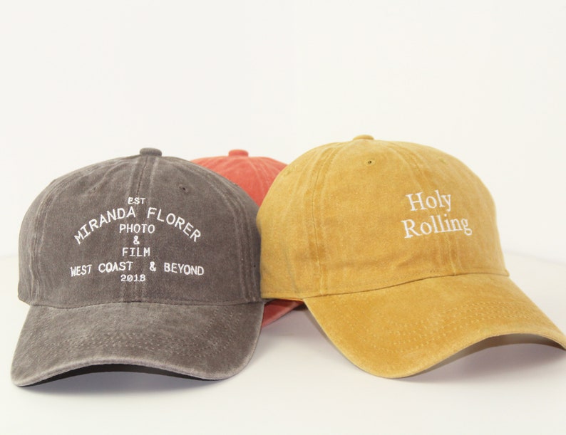 May include: Three baseball caps, one grey with the text "Miranda Florer Est. Photo & Film West Coast & Beyond 2018", one orange, and one yellow with the text "Holy Rolling" in white.