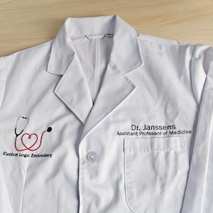 Puede incluir: Bata de laboratorio blanca con cuello, bolsillo y cierre de botón. Cuenta con bordado de logotipo personalizado de un estetoscopio rojo y negro con forma de corazón. El texto "Dr. Janssens, Assistant Professor of Medicine" está impreso.