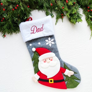 May include: A gray Christmas stocking with a white cuff embroidered with the word "Dad" in red. The stocking features a Santa Claus applique and a white snowflake. Green pine branches with red berries are in the background.