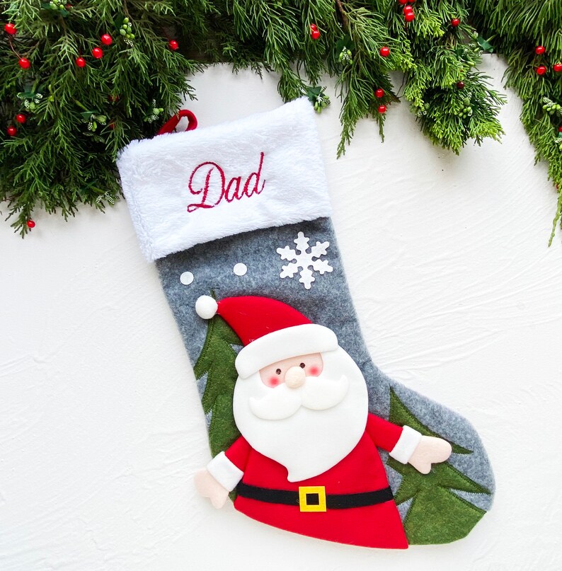 May include: A gray Christmas stocking with a white cuff embroidered with the word "Dad" in red. The stocking features a Santa Claus applique and a white snowflake. Green pine branches with red berries are in the background.