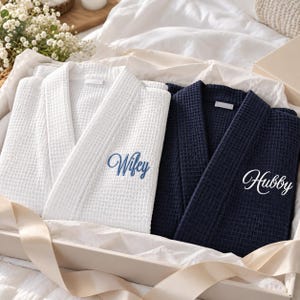 Puede incluir: Una bata blanca de tejido de gofre con el texto bordado en azul Wifey y una bata azul marino de tejido de gofre con el texto bordado en blanco Hubby. Ambas batas tienen cuello de chal y están dobladas cuidadosamente en una caja de regalo.