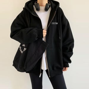 Op de afbeelding: Zwarte hoodie met rits en een witte capuchon. De hoodie heeft het woord "CUSTOM" op de voorkant en de woorden "Life's too short to be anything but" op de achterkant gedrukt.