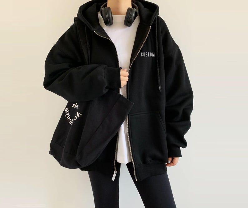 Op de afbeelding: Zwarte hoodie met rits en een witte capuchon. De hoodie heeft het woord "CUSTOM" op de voorkant en de woorden "Life's too short to be anything but" op de achterkant gedrukt.