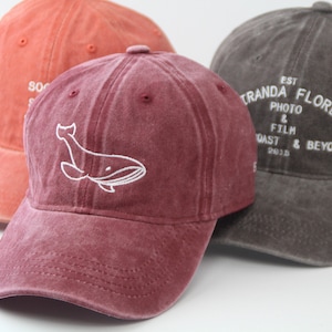 May include: Three baseball caps, one red, one burgundy with a white whale design, and one grey with white text that reads "EST. 2018 TRANDA FLORES PHOTO & FILM COAST & BEYOND".