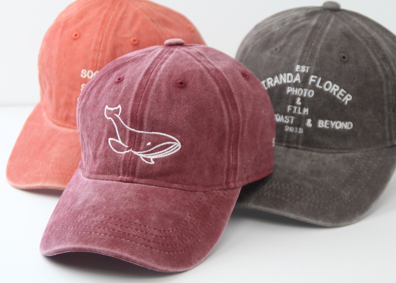 May include: Three baseball caps, one red, one burgundy with a white whale design, and one grey with white text that reads "EST. 2018 TRANDA FLORES PHOTO & FILM COAST & BEYOND".
