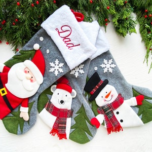 May include: Christmas stockings with festive designs. One features Santa Claus, another a polar bear, and the third a snowman. The stockings are gray with white accents and snowflakes. The top stocking has the word "Dad" embroidered.