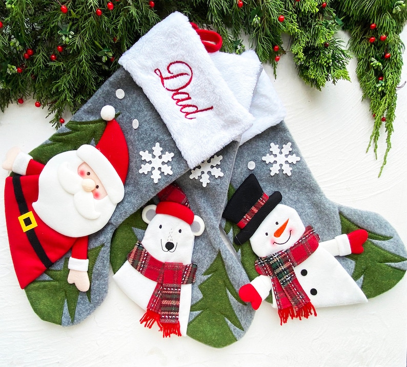 May include: Christmas stockings with festive designs. One features Santa Claus, another a polar bear, and the third a snowman. The stockings are gray with white accents and snowflakes. The top stocking has the word "Dad" embroidered.