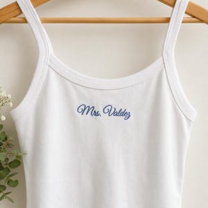 Op de afbeelding: Een wit hemdje met dunne schouderbandjes en een ronde halslijn. In het midden van de borst staat de tekst Mrs. Valdez geborduurd in een blauw cursief lettertype. Het kledingstuk hangt aan een houten kleerhanger tegen een effen achtergrond.