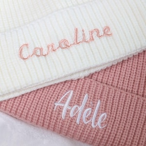 May include: Two knitted beanies, one white with the name "Caroline" embroidered in pink, and one pink with the name "Adele" embroidered in white.