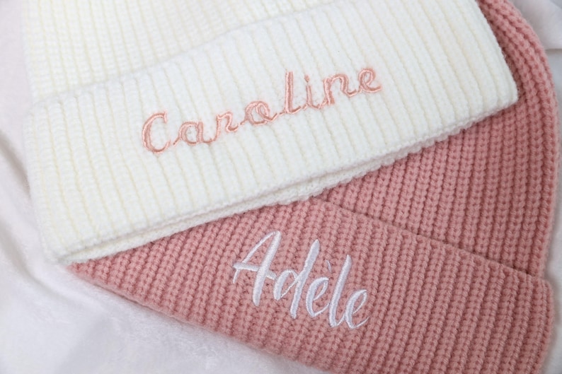 May include: Two knitted beanies, one white with the name "Caroline" embroidered in pink, and one pink with the name "Adele" embroidered in white.