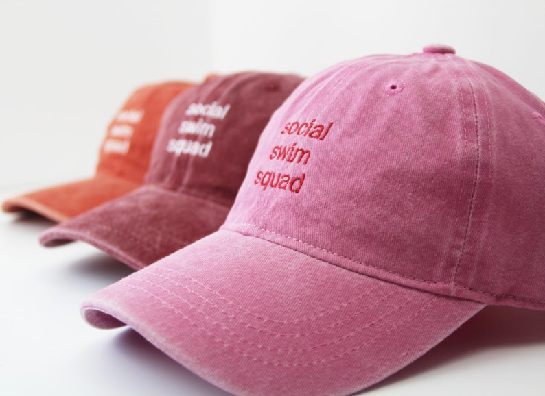May include: Three baseball caps in different shades of pink and red. The caps have the text "social swim squad" embroidered on the front in white.