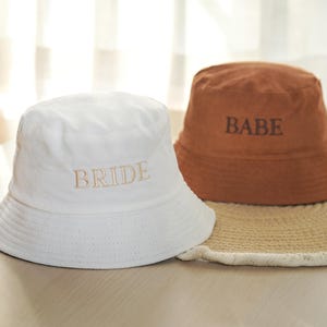 May include: Two bucket hats, one white and one brown, are displayed. The white hat has "BRIDE" embroidered in a light tan color. The brown hat has "BABE" embroidered in a darker brown. Both hats are sitting on a woven mat.