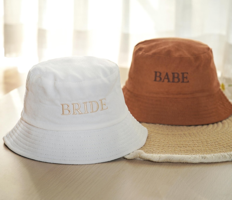 May include: Two bucket hats, one white and one brown, are displayed. The white hat has "BRIDE" embroidered in a light tan color. The brown hat has "BABE" embroidered in a darker brown. Both hats are sitting on a woven mat.