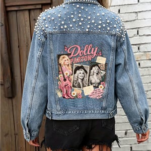 May include: A blue denim jacket featuring white pearl embellishments across the shoulders and back. The back displays a graphic print of Dolly Parton with the text Dolly Parton, a guitar, cowboy boots, flowers, and butterflies in pink and brown.