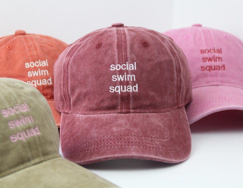 May include: Three baseball caps in different colours: red, pink, and green. Each cap has the text "social swim squad" embroidered on the front in white.