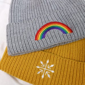 May include: Two knitted beanies, one gray with a rainbow embroidered on it and one yellow with a white snowflake embroidered on it.