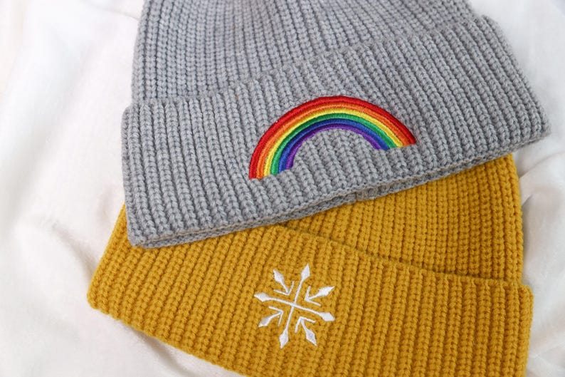 May include: Two knitted beanies, one gray with a rainbow embroidered on it and one yellow with a white snowflake embroidered on it.