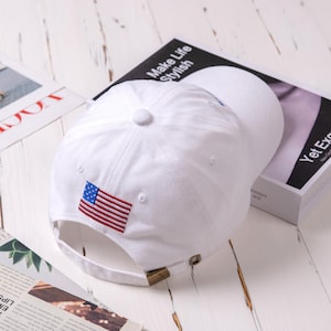 May include: White baseball cap with an embroidered American flag on the front. The cap has an adjustable strap in the back.