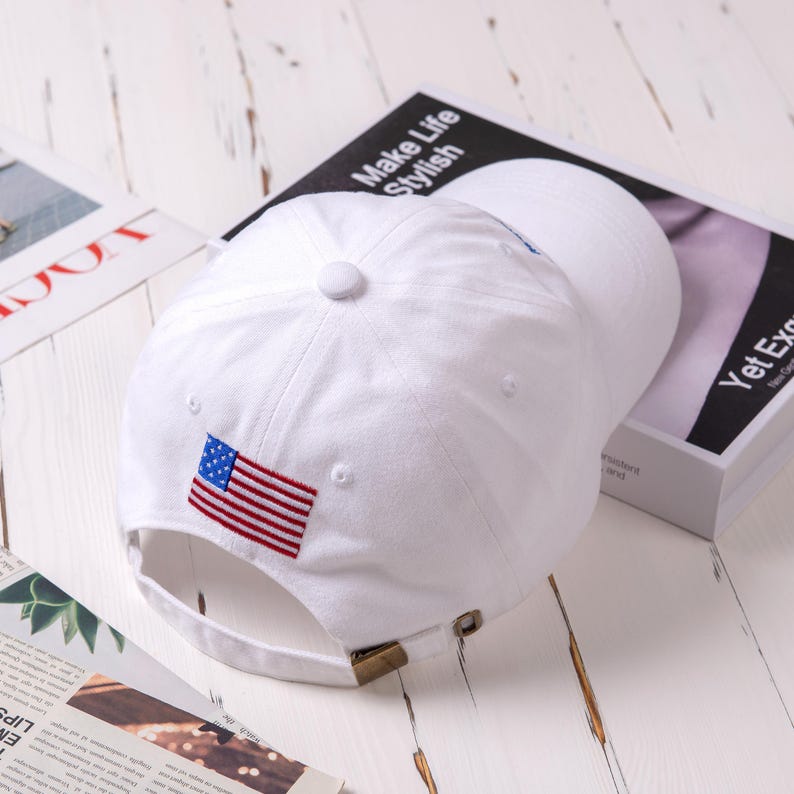 May include: White baseball cap with an embroidered American flag on the front. The cap has an adjustable strap in the back.