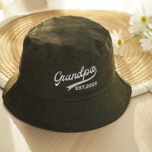 May include: An olive green bucket hat with the word "Grandpa" embroidered in white script, along with "EST.2025". The hat is made of a textured fabric and has a wide brim. The hat is resting on a straw hat.