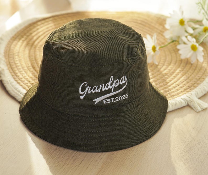 May include: An olive green bucket hat with the word "Grandpa" embroidered in white script, along with "EST.2025". The hat is made of a textured fabric and has a wide brim. The hat is resting on a straw hat.