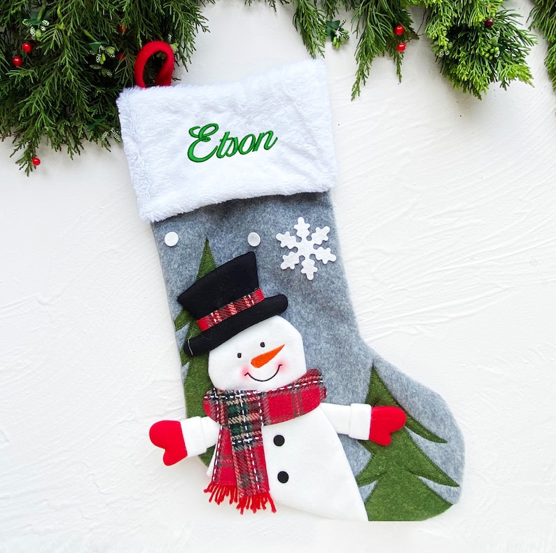May include: A gray Christmas stocking with a white cuff and a red loop. The stocking features a snowman with a black top hat, red scarf, and red mittens. The name "Etson" is embroidered in green on the cuff. A white snowflake and green trees are also on the stocking.