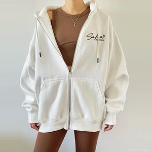 Op de afbeelding: Witte hoodie met rits en een zwart geborduurd logo met de tekst "Sofia" en "Be Inspired".