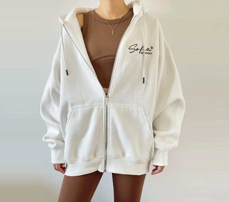 Op de afbeelding: Witte hoodie met rits en een zwart geborduurd logo met de tekst "Sofia" en "Be Inspired".