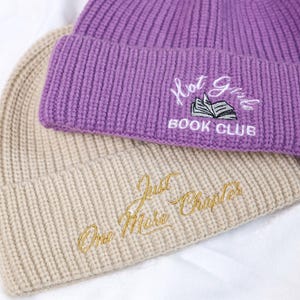 May include: Two knitted beanies, one beige and one purple. The beige beanie has the text "One More Chapter" embroidered on it in gold thread. The purple beanie has the text "Hot Girls Book Club" embroidered on it in white thread.