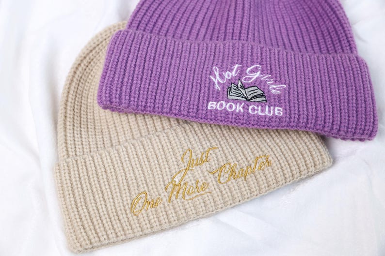 May include: Two knitted beanies, one beige and one purple. The beige beanie has the text "One More Chapter" embroidered on it in gold thread. The purple beanie has the text "Hot Girls Book Club" embroidered on it in white thread.