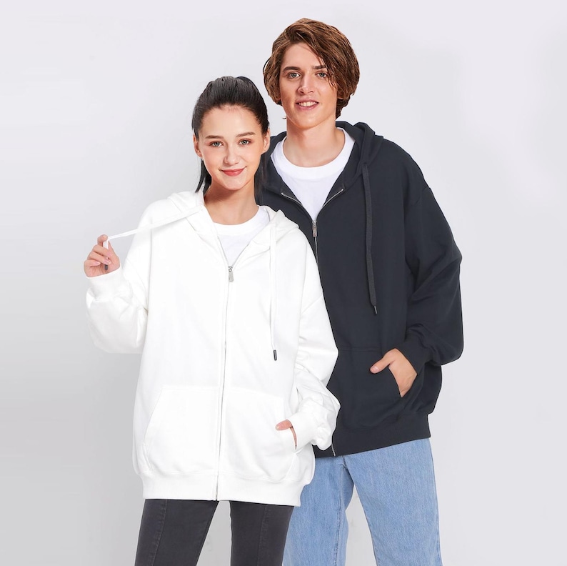 May include: Two people wearing hooded sweatshirts, one white and one black. Both sweatshirts have zipper closures.
