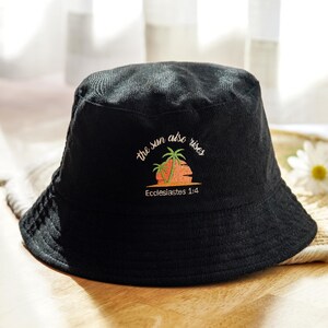 May include: Black corduroy bucket hat with embroidered text "the sun also rises" above a sunset and palm tree design. The text "Ecclesiastes 1:4" is below the design. The hat is sitting on a woven surface.