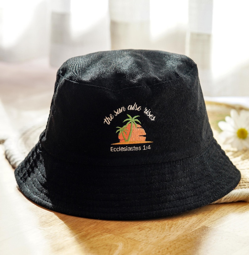 May include: Black corduroy bucket hat with embroidered text "the sun also rises" above a sunset and palm tree design. The text "Ecclesiastes 1:4" is below the design. The hat is sitting on a woven surface.
