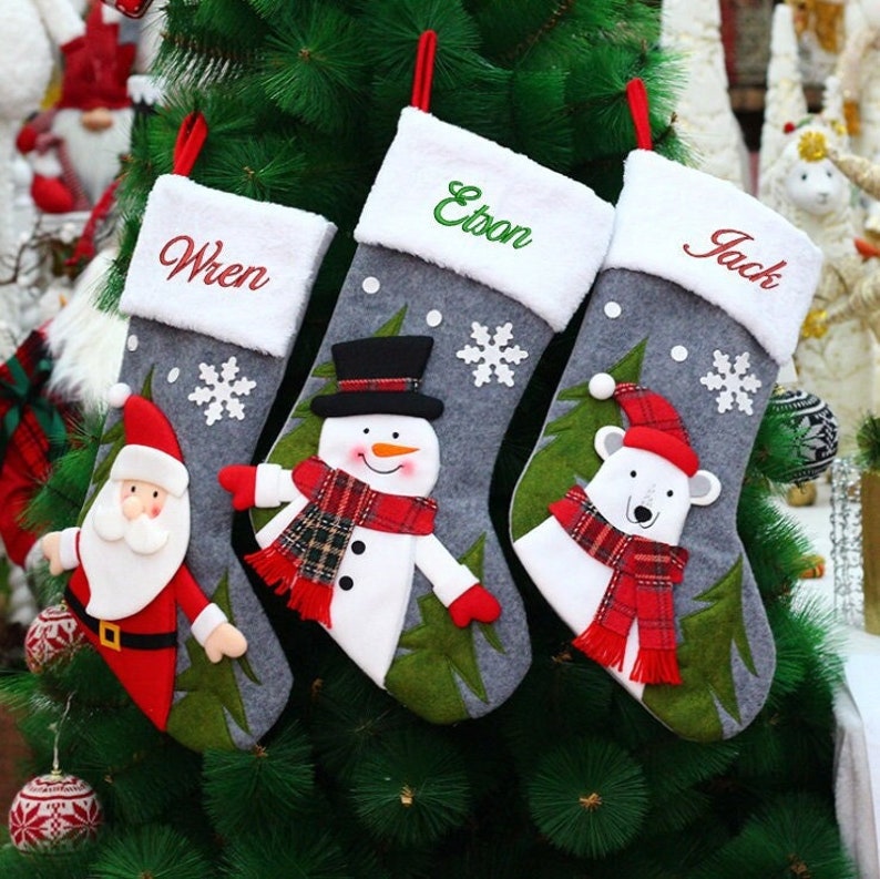 May include: Three gray Christmas stockings with white cuffs hang on a green tree. Each stocking has a different design: Santa Claus, a snowman, and a polar bear. The names "Wren", "Etson", and "Jack" are embroidered on the cuffs.