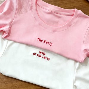 Puede incluir: Dos camisetas apiladas, una rosa y otra blanca. La camiseta rosa tiene el texto bordado en rojo "The Party". La camiseta blanca tiene el texto bordado en rojo "Wife of the Party". Ambas camisetas tienen cuello redondo.