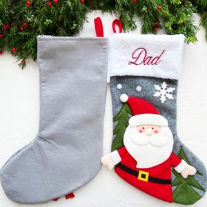 May include: Two Christmas stockings. One is solid gray, the other is gray with a Santa Claus design. The Santa stocking has a red hat and coat, white beard, and a gold belt buckle. The word "Dad" is embroidered in red on the white cuff.