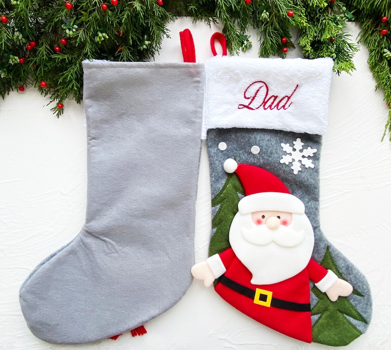 May include: Two Christmas stockings. One is solid gray, the other is gray with a Santa Claus design. The Santa stocking has a red hat and coat, white beard, and a gold belt buckle. The word "Dad" is embroidered in red on the white cuff.