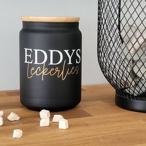 Könnte beinhalten: Schwarze Keramikdose mit Holzdeckel, beschriftet mit "EDDYS Leckerlies". Die Dose ist mit kleinen weißen Leckerlis gefüllt und steht auf einer Holzoberfläche.