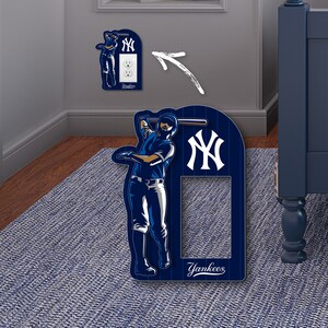 May include: A blue and white New York Yankees light switch cover with a baseball player holding a bat. The cover has the team logo and the word "Yankees" on it.