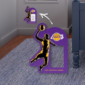 May include: A purple and gold Los Angeles Lakers basketball player wall decal. The decal is shaped like a basketball player in mid-air, holding a basketball. The decal is designed to be placed over a light switch.