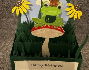 Könnte beinhalten: Handgefertigte Geburtstagskarte mit einem grünen Frosch mit goldener Krone, der auf einem rot-weißen Pilz sitzt. Die Karte hat einen blauen Himmel, gelbe Blumen und grünes Gras. Die Worte "Happy Birthday" sind auf der Vorderseite zu sehen.