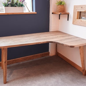 May include: A corner desk made of reclaimed wood with a natural finish. The desk has a curved front edge and is supported by four legs. The desk is in a room with a light blue wall and a white wall.